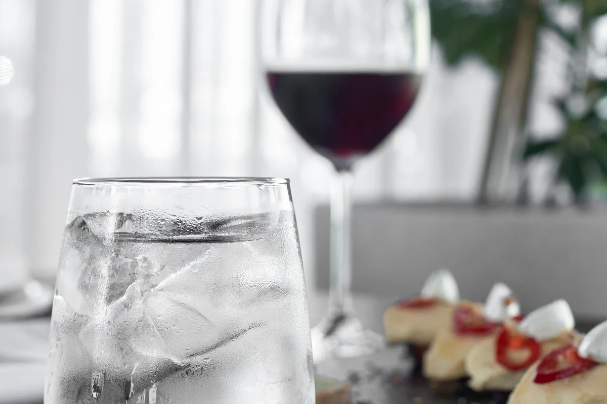 A glass of red wine beside iced water and food at a dining table.